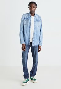 Camisa de mezclilla azul claro sobre una camiseta blanca, combinada con jeans azul oscuro y zapatillas verdes. Presenta bolsillos clásicos con botones y un corte relajado.