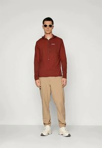 Punane pika varrukaga pullover pool lukuga, kantud beežide pükste kohal. Modellil on päikeseprillid ja valged tossud.