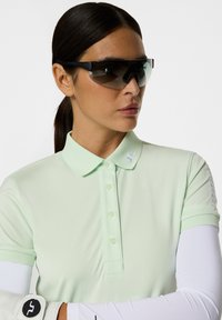 Vaaleanvihreä polo-paita, jossa on kaulus ja kolme nappia, yhdistettynä mustiin urheilullisiin aurinkolasikohteisiin ja valkoiseen pitkähiharaiseen aluspaitaan.