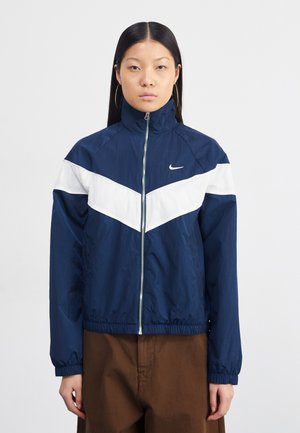 Navy blue windbreaker jacket with white chevron pattern, high collar, full zipper, elastic cuffs, and waistband. Made from lightweight, textured fabric.