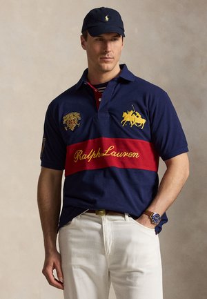 Άνδρας που φοράει μπλε σκούφο και μπλε πόλο με κόκκινη ρίγα, κίτρινο λογότυπο παίκτη πόλο και άσπρο παντελόνι, σε απλό φόντο.