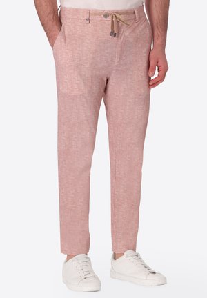 Uomo che indossa pantaloni di lino rosa chiaro con coulisse beige, tasche laterali e sneakers bianche, in piedi davanti a uno sfondo semplice.