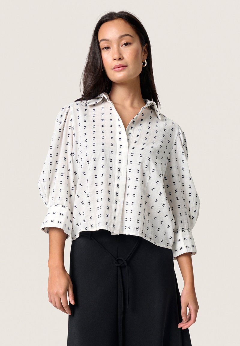 Chemise blanche avec un motif croix noir, col boutonné, épaules tombantes et manches trois-quarts. Tissu léger, design court.