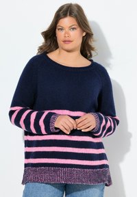 Vrouw met golvend bruin haar in een marineblauwe trui met roze strepen, staand tegen een witte achtergrond.