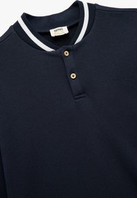 Suéter de algodón azul marino con cuello redondeado, acentos a rayas blancas y dos botones de madera en el broche. Textura suave y lisa.