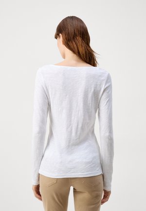 Femme aux longs cheveux bruns portant une chemise blanche à manches longues et un pantalon beige, vue de dos sur un fond uni.