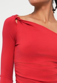 Czerwona bluzka z długim rękawem i skręconym detalem na ramieniu. Gładki, elastyczny materiał z bocznymi marszczeniami dla dopasowanego sylwetki.