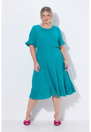 Donna in vestito color teal lungo fino al ginocchio con maniche e orlo arricciati, indossa tacchi rosa aperti con fiocco, in piedi su sfondo bianco.