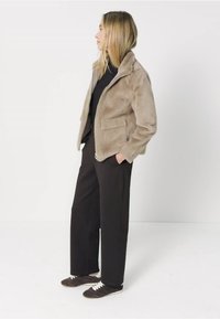 Vrouw staat zijwaarts, gekleed in een beige nepbontjack, zwarte broek, zwart topje en zwarte sneakers met witte veters, tegen een eenvoudige witte achtergrond.