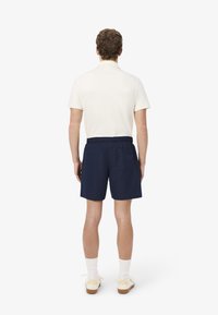 Costume da bagno della marina con vita elastica e tasca sul retro, realizzato in materiale leggero. Indossato con una polo crema e calzini bianchi.