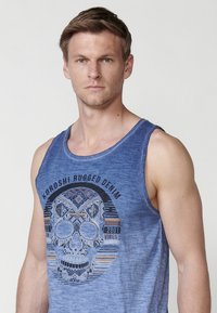 Kék ujjatlan póló puha anyagból, grafikus koponya mintával, geometrikus mintákkal és "KOROSHI RUGGED DENIM 2001 VIBES" felirattal.