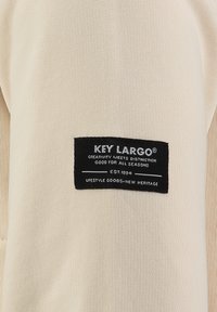 T-shirt beige con un'etichetta in tessuto nera riportante testo bianco, che indica "KEY LARGO" e altri dettagli del marchio. Design a maglia testurizzata.