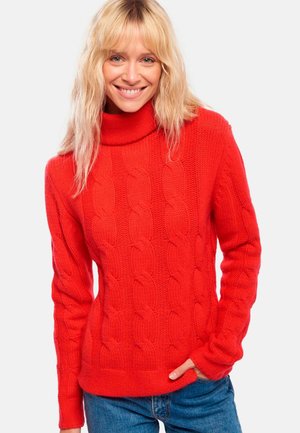Pull en maille rouge avec un col montant, présentant un motif en torsades et des poignets côtelés. Associé à un jean en denim bleu.