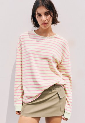 Jeune femme aux cheveux courts et foncés porte une chemise à manches longues rayée jaune pâle et rose et une minijupe beige avec des poches.
