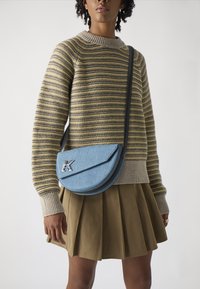 Borsa a tracolla in denim azzurro chiaro con forma arrotondata, rifiniture nere e un accento in metallo argentato a forma di lettera "K"; indossata con un maglione a righe.