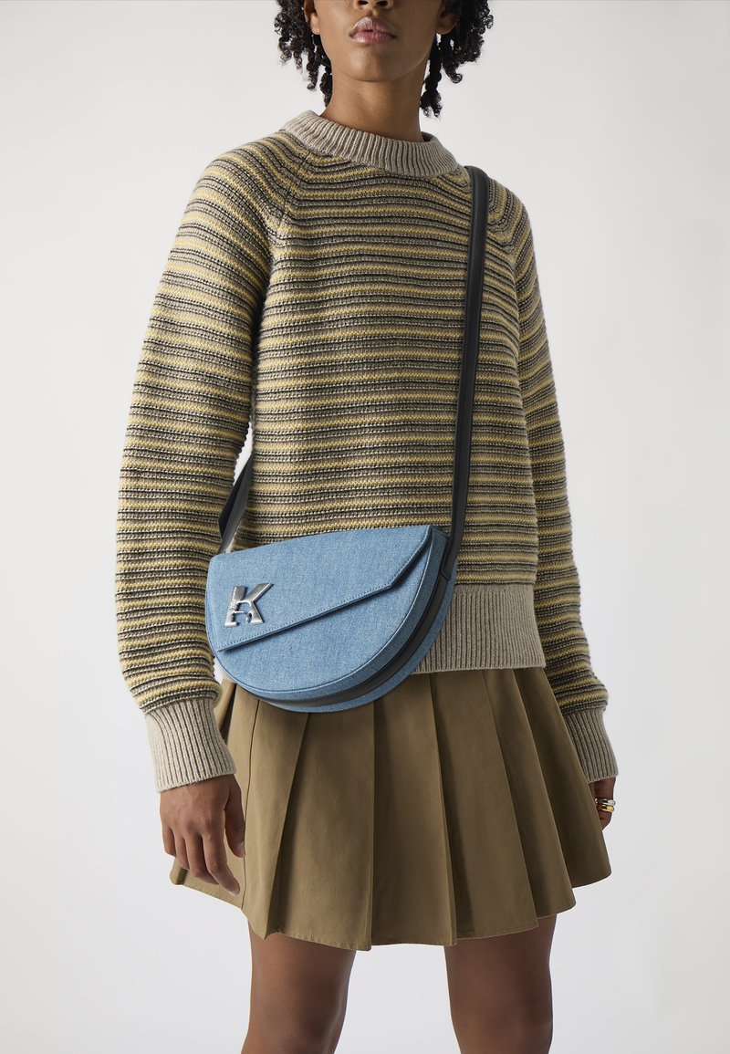Borsa a tracolla in denim azzurro chiaro con forma arrotondata, rifiniture nere e un accento in metallo argentato a forma di lettera "K"; indossata con un maglione a righe.