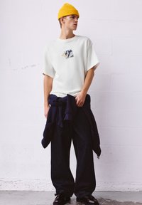 Weißes, locker sitzendes T-Shirt mit grafischem Design, kombiniert mit dunklen, weiten Jeans und einem marineblauen Sweatshirt, das um die Taille gebunden ist. Gelbe, gerippte Mütze.