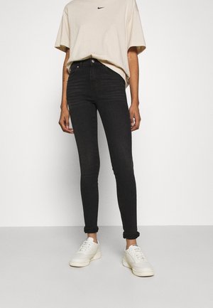 Osoba ubrana w beżową koszulkę Nike, czarne obcisłe dżinsy z wysokim stanem i podwiniętymi nogawkami oraz białe trampki, stojąca na jednolitym białym tle.