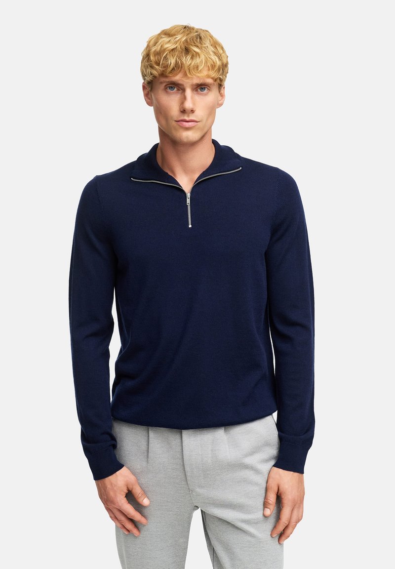 Maglione zip-up blu navy in un materiale morbido e testurizzato. Presenta un colletto a imbuto e maniche lunghe, abbinato a pantaloni grigi chiari.