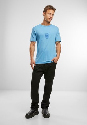 Himmelblaues Baumwoll-T-Shirt mit Rundhalsausschnitt und kurzen Ärmeln, mit einem dunkelblauen Emblem auf der Brust. Kombiniert mit schwarzen Jeans und schwarzen Stiefeln.