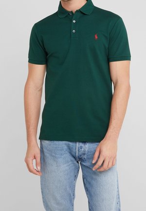 Homme portant un polo vert foncé avec un logo rouge et un jean bleu clair, debout devant un fond gris uni.