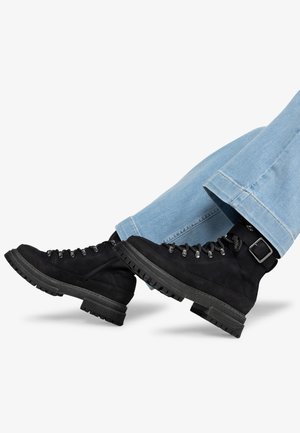 Bottes de randonnée en daim noir avec des semelles en caoutchouc épaisses, œillets en métal et boucles décoratives, associées à un pantalon en denim bleu clair.