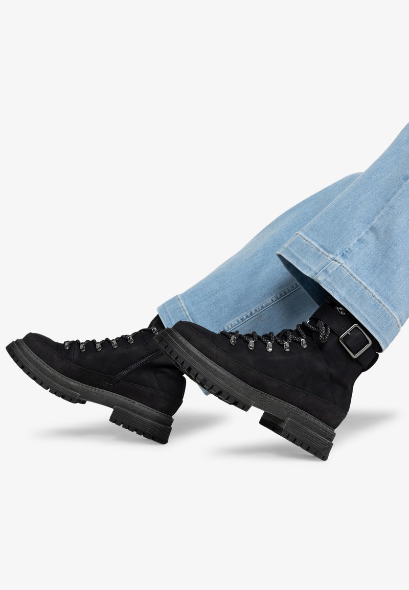 Stivali da escursionismo in suede nero con suole in gomma spesse, occhielli in metallo e fibbie decorative, abbinati a pantaloni in denim azzurro chiaro.