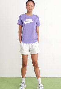 Vijolična bombažna majica s belim Nike logotipom, v kombinaciji z belimi kratkimi hlačami, nevtralnim obutvijo in nogavicami do gležnjev. Preprosta, sproščena športna obleka.