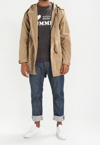 Beige lättviktsjacka med huva, grå grafiskt t-shirt, mörkblå jeans med uppvikta ärmar och vita sneakers. Rena linjer och avslappnad passform.