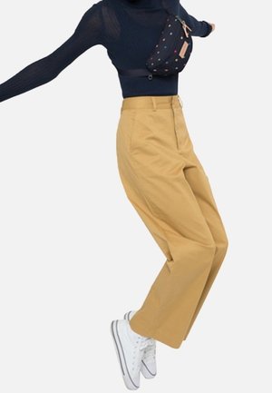 Persona con pantalones anchos de color beige, zapatillas blancas, una camiseta negra de manga larga y un bolso bandolera negro con puntos de colores, capturada en pleno salto.