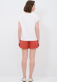 Weißes kurzärmeliges Oberteil mit rundem Ausschnitt, kombiniert mit rostfarbenen Shorts. Der Stoff wirkt glatt, mit klaren Linien und gefalteten Saumdetails.