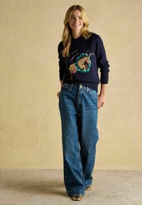 Marineblauer Pullover mit Weihnachtsmotiv, der ein Rentier zeigt; kombiniert mit weit geschnittenen blauen Jeans. Goldene Schuhe runden das Outfit ab.