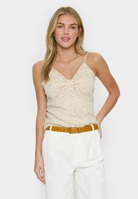 Camisola in pizzo beige con scollatura a V e spalline sottili, caratterizzata da un dettaglio arricciato davanti, indossata con pantaloni bianchi a vita alta e una cintura marrone.