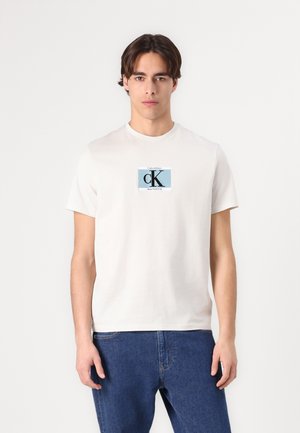 Camiseta de algodón en color crema con cuello redondo, que presenta un logotipo gráfico azul en la parte delantera. Se lleva con unos vaqueros de mezclilla azules.