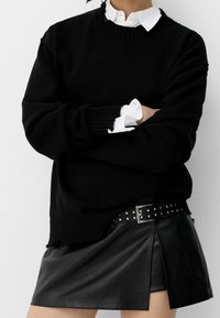 Suéter negro de punto con cuello y puños acanalados, utilizado sobre una camisa blanca con collar. Combinado con una mini falda de cuero negra y un cinturón con tachuelas.