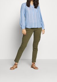 Blusa a maniche lunghe a righe blu chiaro con chiusura a bottoni, abbinata a pantaloni cargo verde oliva e sandali marroni con cinturini.