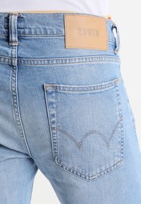Ljusblå denimjeans med en läderetikett märkt "EDWIN." Har dubbla sömmar och en distinkt böjd fickdesign.