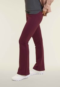 Pantaloni a palazzo bordeaux realizzati in tessuto elasticizzato, con cuciture laterali e una finitura liscia. Indossati con scarpe da ginnastica bianche.