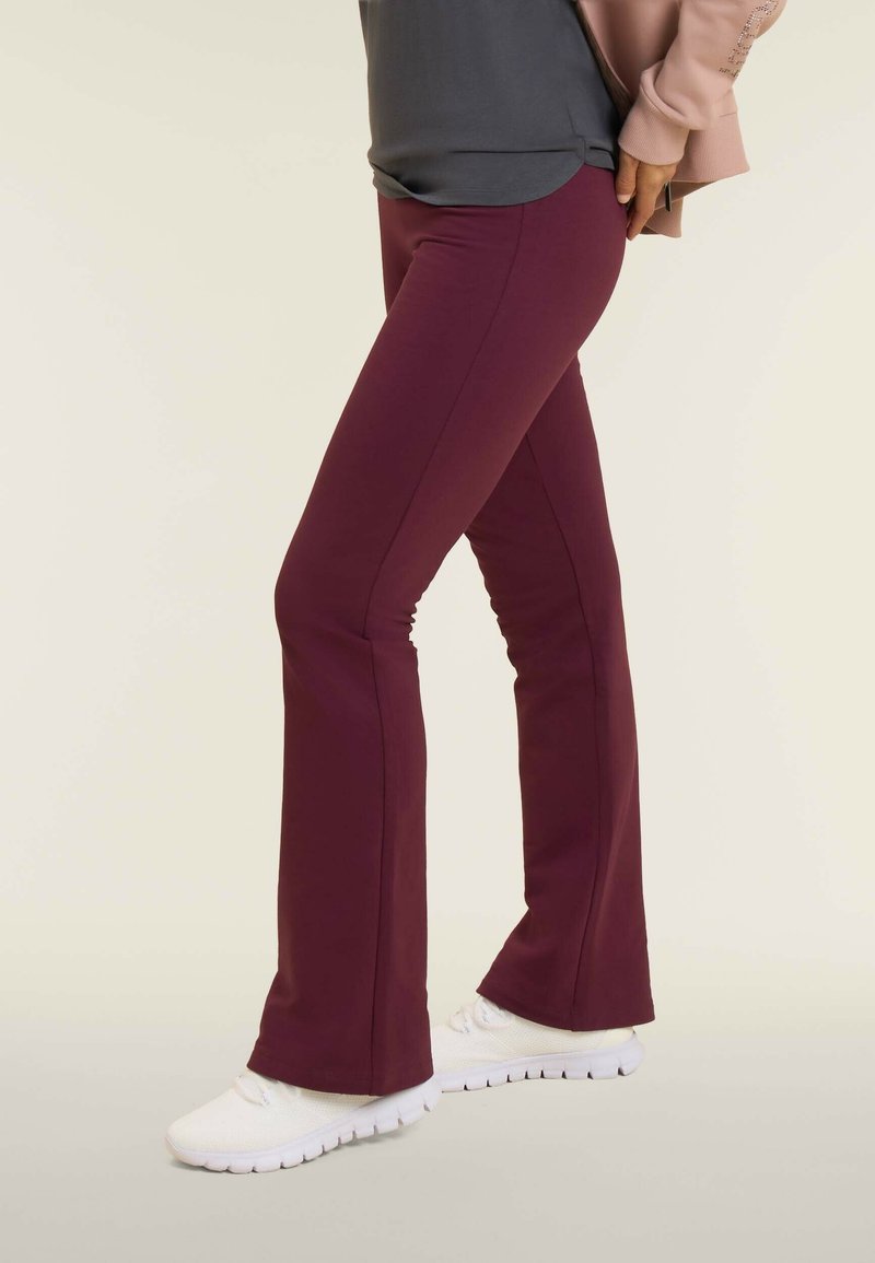 Pantaloni a palazzo bordeaux realizzati in tessuto elasticizzato, con cuciture laterali e una finitura liscia. Indossati con scarpe da ginnastica bianche.