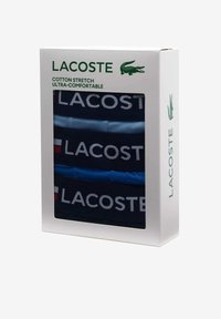 Chaussettes en coton extensible dans un ensemble de trois, affichant des couleurs bleu marine et bleu clair avec un grand logo blanc 'LACOSTE'. Présentées dans une boîte blanche.