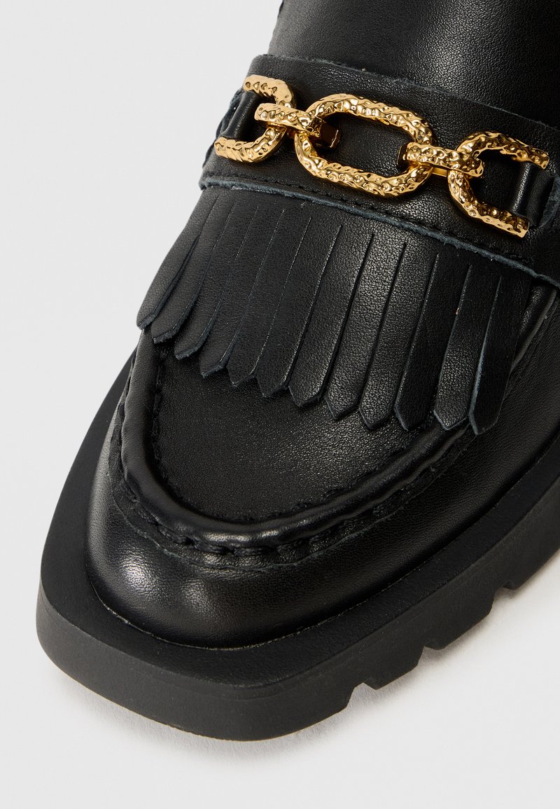 Schwarzer Lederschuh mit fransigem Detail und einer goldenen strukturierten Kette. Verfügt über eine abgerundete Zehe und eine dicke Gummisohle.