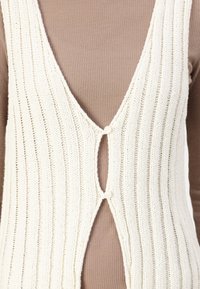 Strickweste in Creme mit vertikalen Rippungen entlang der Kanten, mit einem tiefen V-Ausschnitt und einer Knopfleiste in der Mitte vorne.