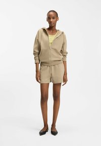 Frau trägt eine beige Jacke mit Reißverschluss und Shorts, darunter ein hellgrünes Oberteil, sowie schwarze flache Schuhe, steht vor einem weißen Hintergrund.