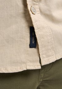 Lichtbeige linnen overhemd met een knoopdetail en een donkerblauw merklabel. De stof heeft een gestructureerde afwerking, in combinatie met olijfgroene broeken.