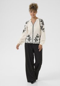 Broderet cremebluse med sorte blomster mønstre, V-hals og elastiske manchetter, parret med sorte brede bukser med subtile udsmykninger.