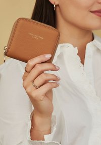 Braunes, genarbtes Lederportemonnaie mit goldenen Beschlägen, Reißverschluss und geprägtem Markenlogo. In einer Hand gehalten, die mit Ringen geschmückt ist. Weißes Blusenshirt mit Spitzenakzenten.