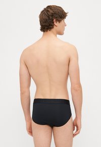Černé plavky s texturovaným pasem s logem, s přiléhavým střihem a hladkým materiálem. Model je viděn zezadu.