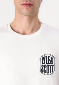 T-shirt de algodão branca com decote redondo e um emblema bordado azul-marinho com "LYLE & SCOTT". Textura suave, ajuste padrão.