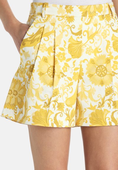 Shorts évasés avec un motif floral jaune sur tissu blanc, poche latérale avec main à l'intérieur, devant plissé, portés avec un haut blanc.