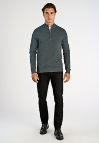 Dunkelgrüner, Reißverschluss-Pullover mit strukturierter Strickart, kombiniert mit schwarzen, schmal geschnittenen Hosen und glänzenden schwarzen Schuhen. Schlichtes Design, keine Muster.